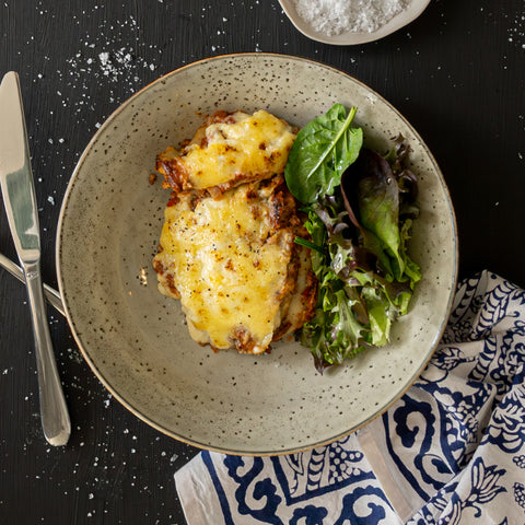 Plated dish of cheesy meat with a side of greens on a speckled plate, set on a dark surface with a patterned cloth.