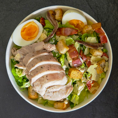 Salad with chicken, eggs, bacon, and croutons in a white bowl on a dark surface