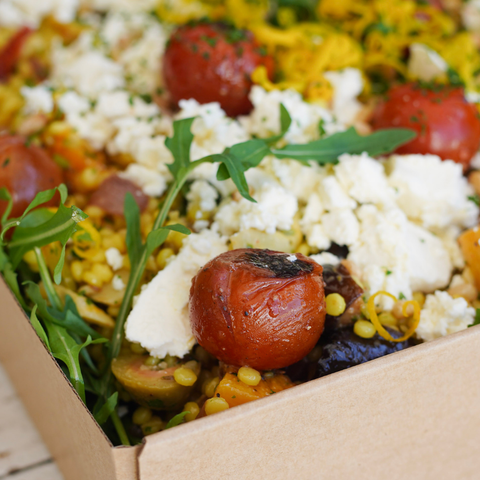 Close-up of a salad with tomatoes, feta cheese, and greens in a cardboard box.