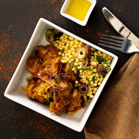 White square bowl with fried dish and couscous on a dark surface with spices and cutlery.