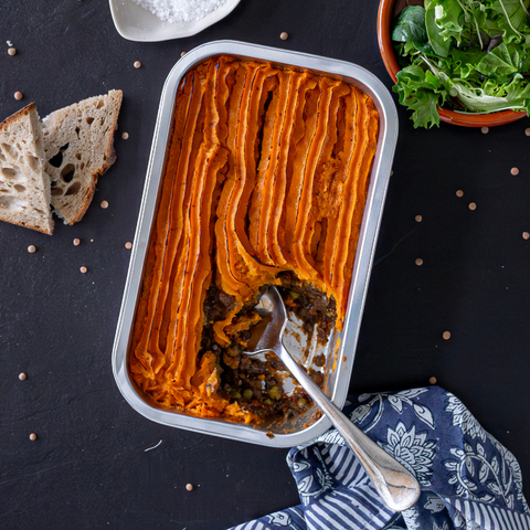 Shepherd's pie in a baking dish with a spoon, surrounded by bread and salad on a dark surface.