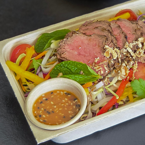 Plated salad with sliced beef, vegetables, and a small bowl of dressing on a dark surface.