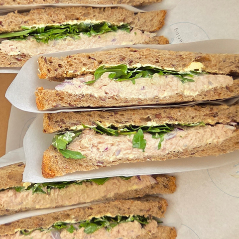 Row of tuna salad sandwiches with arugula on whole grain bread, stacked on a white tray.